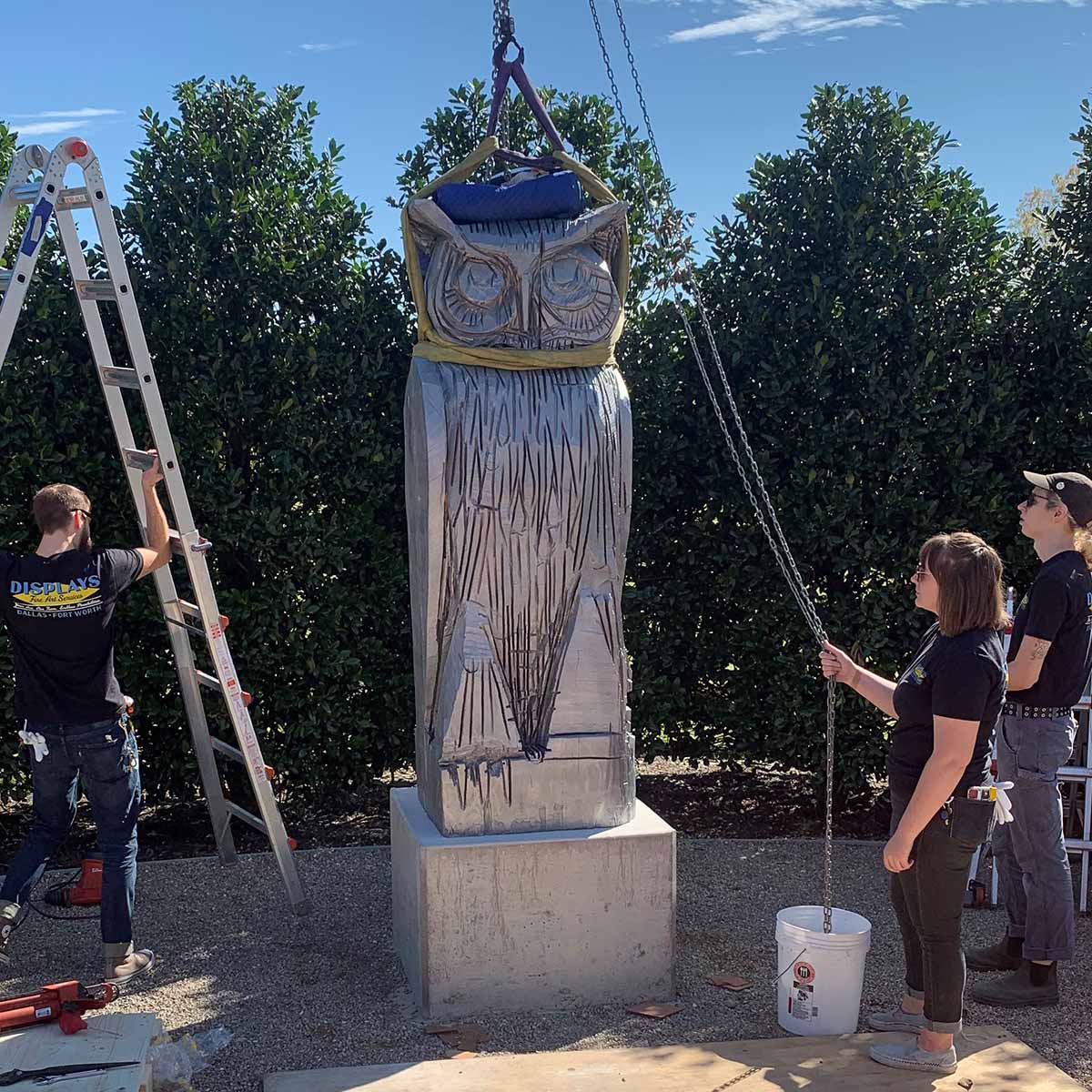 Three Displays Fine Art Services team members install a large statue of an owl outdoors.